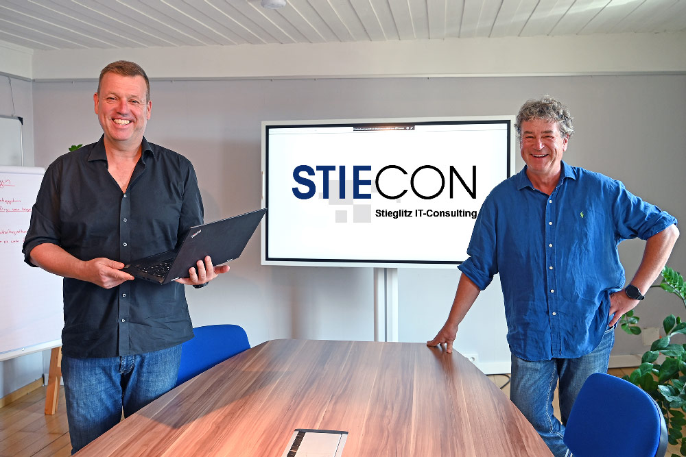 Zwei Mitarbeiter stehen im Büro vor einem Bildschirm, auf dem das StieCon Logo zu stehen ist